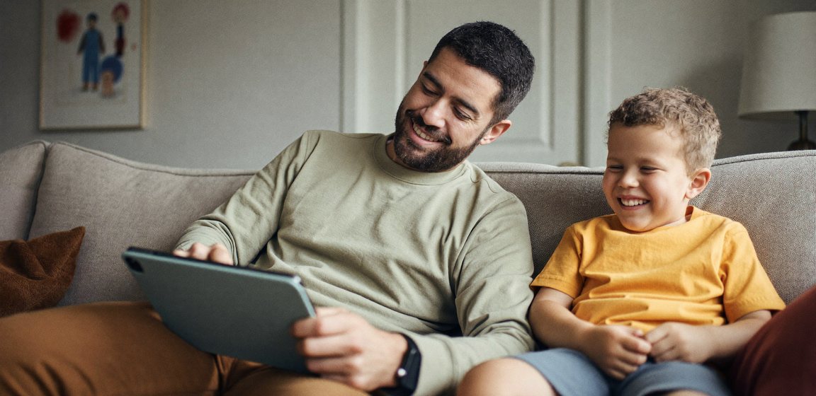 A father and his son sitting together on a couch, looking at a tablet.